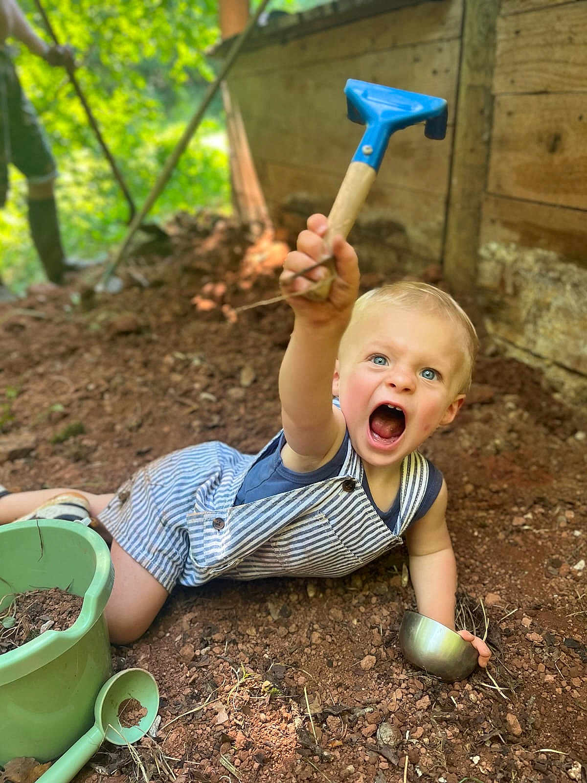 Marcel a rejoint le concours — aidez-le/la à gagner de superbes lots ! toddler, child, blue_eyes, striped_overalls, shovel, metal_bowl, green_bucket, green_scoop, dirt, soil, outdoor, playing, garden, wooden_wall, nature, sunlight, happy, expression, summer, casual_clothing