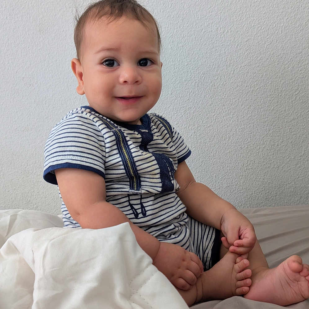 Noah is registered to the contest to win money with this photo: baby, child, infant, smiling, sitting, portrait, indoors, bed, blanket, striped_clothing, short_sleeves, bare_feet, bedroom, wall, cozy, happy, face, person, photography, natural_light