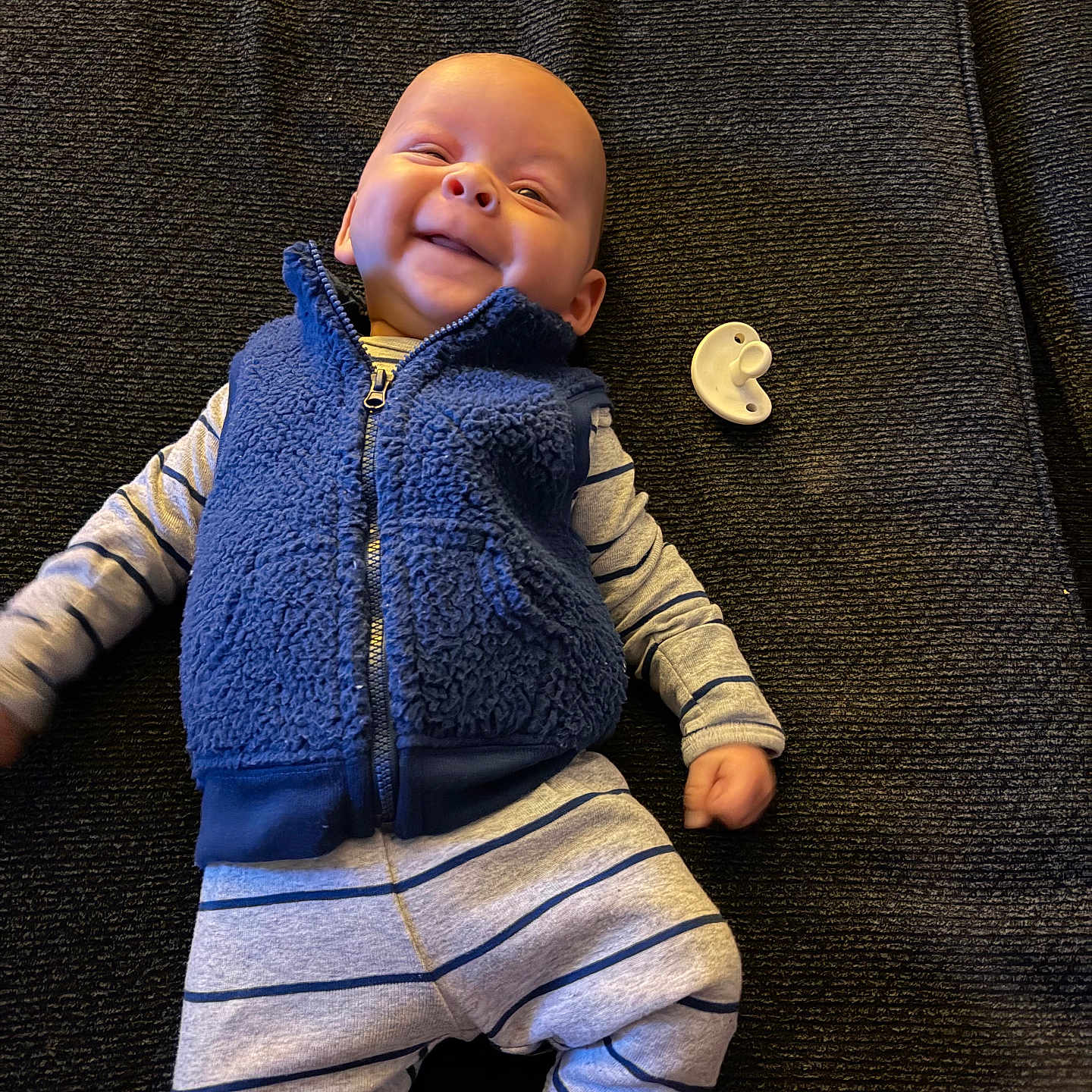 Braxton is registered to the contest to win money with this photo: baby, bedding, blanket, blue_vest, clothing, cute, fabric_texture, face, hand, happy, head, indoor, infant, lying_down, newborn, pacifier, portrait, smile, socks, striped_pajamas