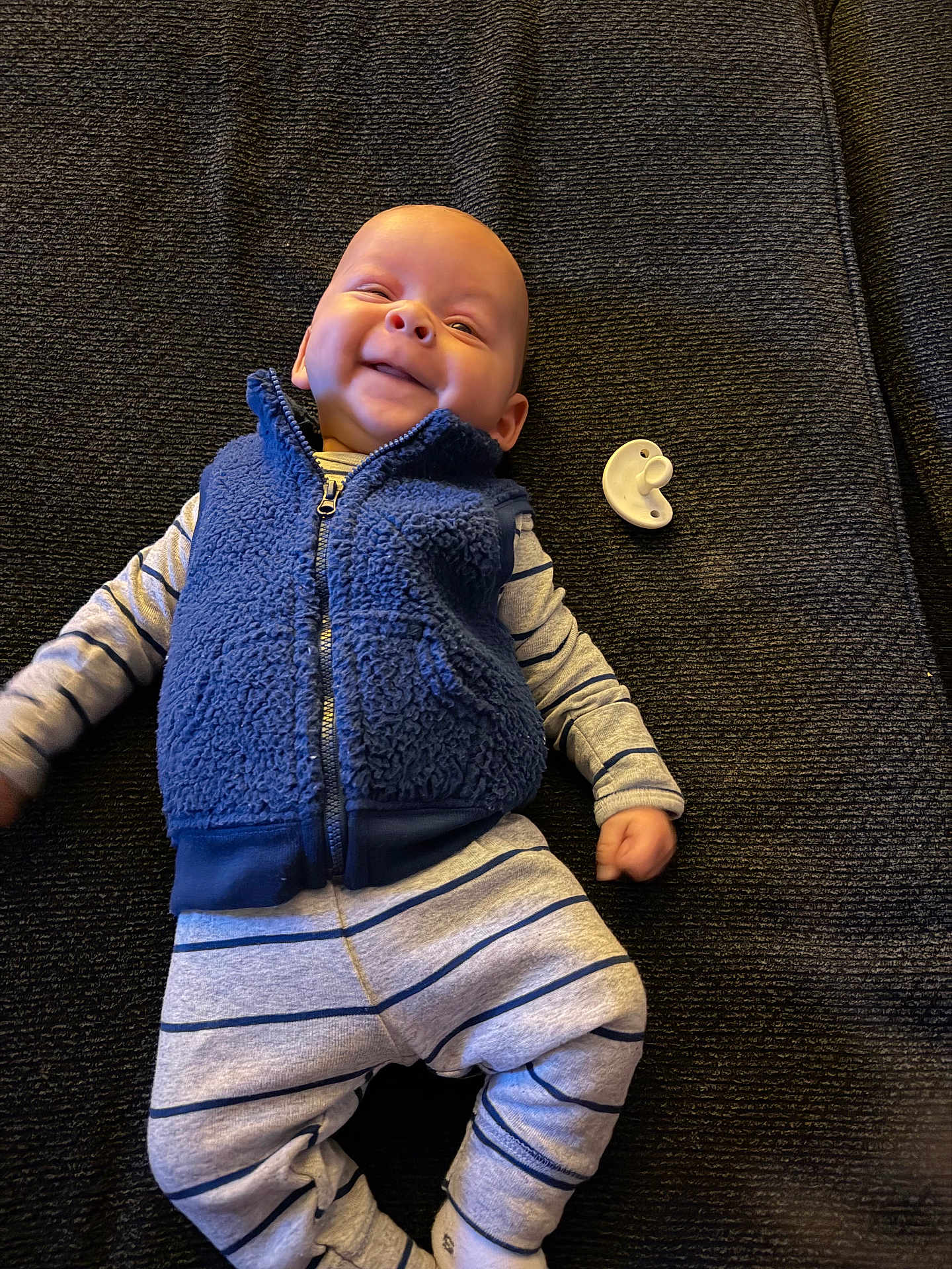 Braxton is registered to the contest to win money with this photo: baby, infant, smile, blue_vest, striped_pajamas, pacifier, blanket, fabric_texture, lying_down, portrait, clothing, happy, face, head, newborn, indoor, bedding, hand, socks, cute