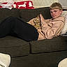 person, cat, couch, blanket, indoor, relaxed, young_person, orange_cat, furniture, home, cozy, patterned_wall, sitting, casual_clothing, hoodie, black_pants, tabby_cat, carpet, table, living_room