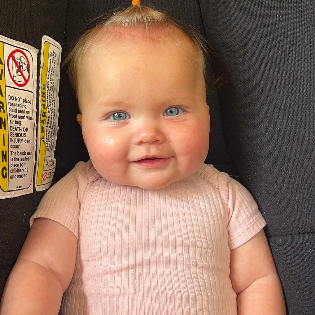 Rivlynn is registered to the contest to win money with this photo: baby, blanket, blue_eyes, car_seat, child, cute, furniture, hand, happy, head, indoor, infant, person, pink_clothing, portrait, seat, skin, smiling, topknot, wood_floor