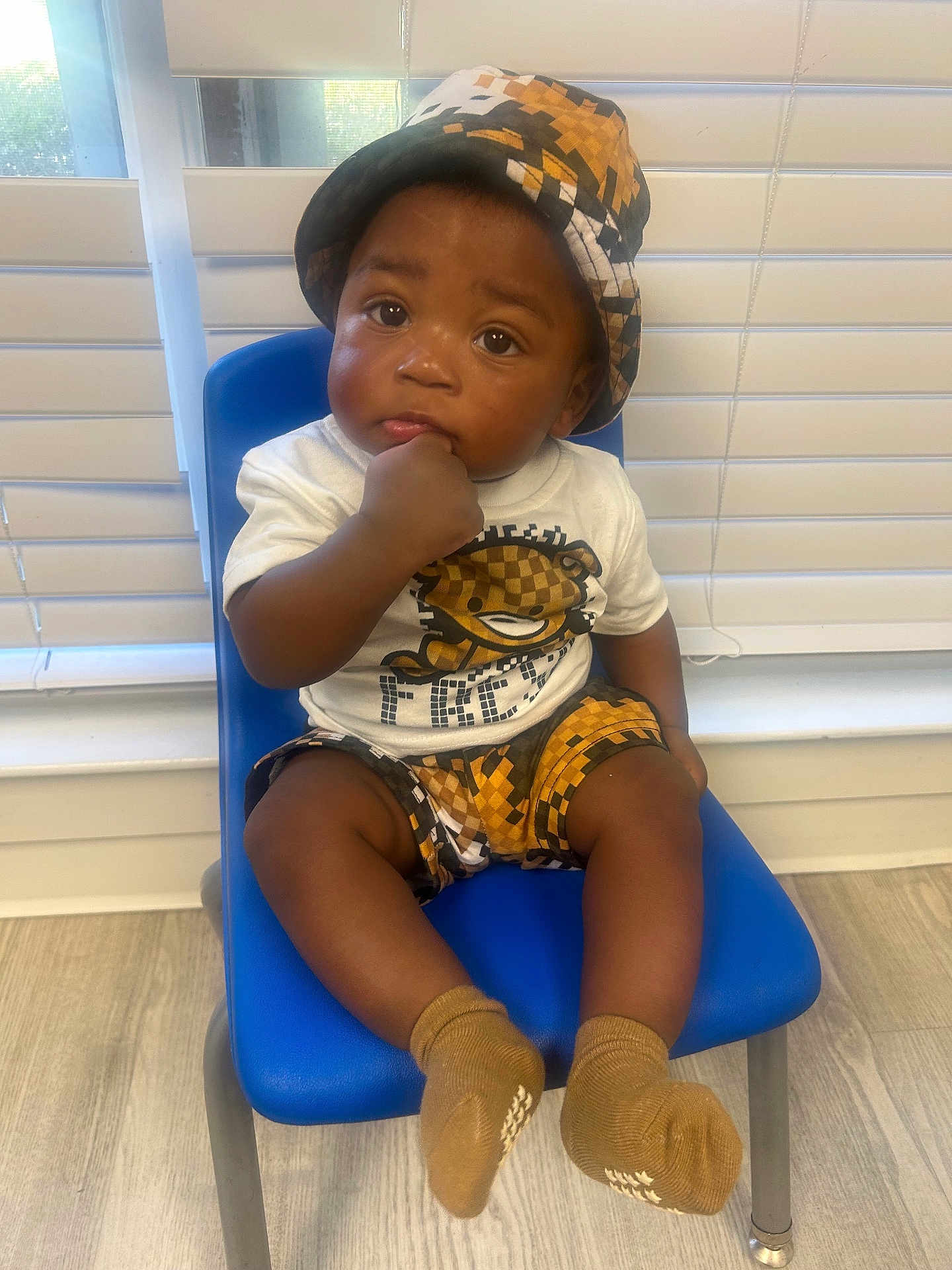 Kaison is registered to the contest to win money with this photo: baby, child, indoor, blue_chair, hat, shorts, socks, tshirt, face, hand, floor, window, blinds, curious, seated, portrait, person, cute, expression, furniture