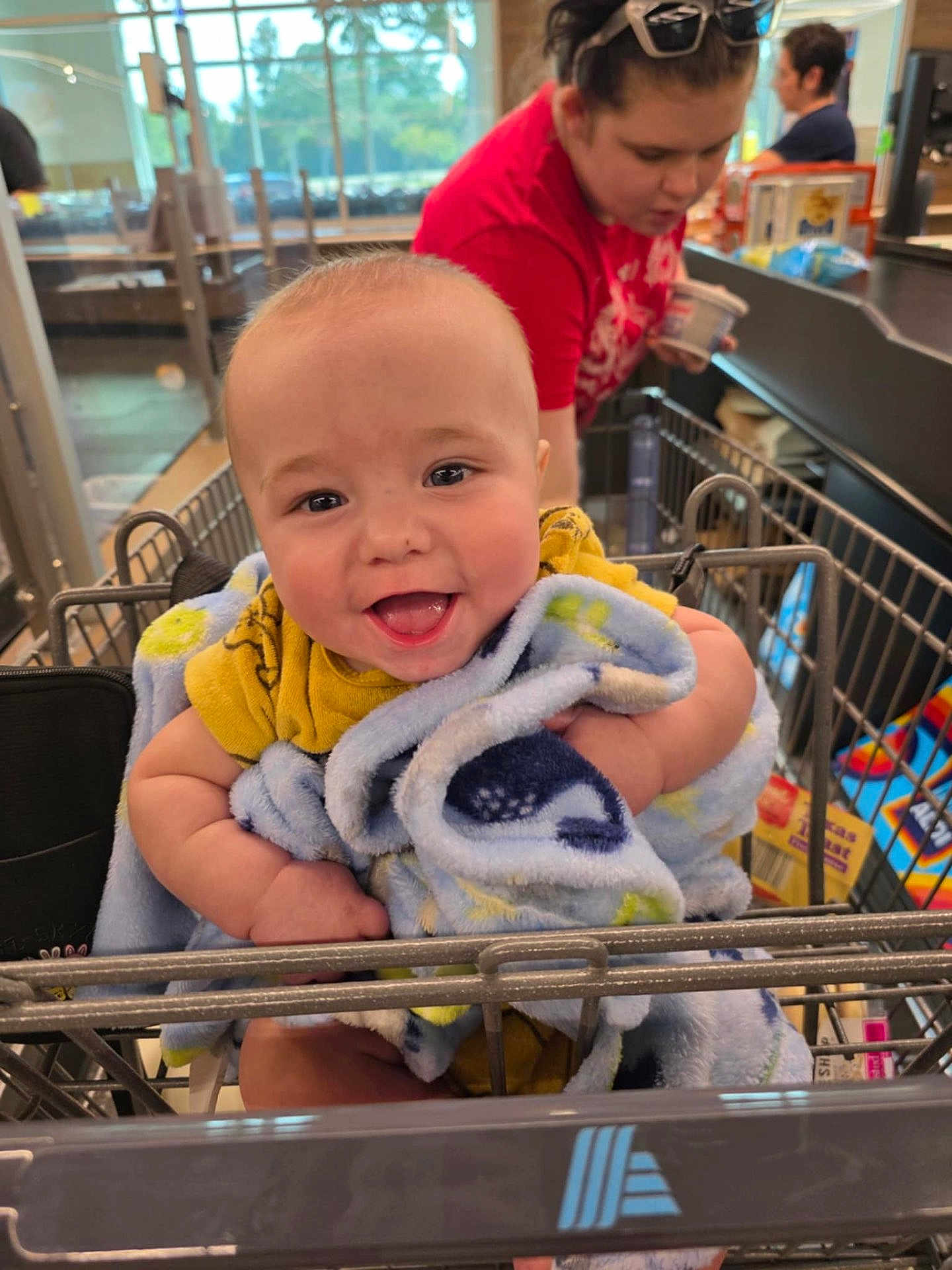 Milo joined the competition — help win amazing prizes! baby, smiling, blanket, shopping_cart, child, indoor, woman, grocery_store, happy, person, retail, clothing, face, young_child, holding, toddler, store, retail_environment, cute, infant