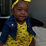 Nova is registered to the contest to win money with this photo: baby, child, infant, smile, yellow_dress, headband, denim_jacket, earrings, cute, happy, portrait, sitting, indoors, soft_blanket, closeup, dark_skin, curly_hair, fashion, adorable, young_child