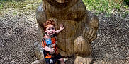 Kaïs a rejoint le concours — aidez-le/la à gagner de superbes lots ! child, toddler, curly_hair, orange_shirt, black_shorts, white_sneakers, water_bottle, wooden_bear, sculpture, forest, trees, leaves, dirt_path, outdoor, nature, daylight, person, pose, playful, wood