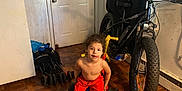 Jayden is registered to the contest to win money with this photo: child, indoor, tricycle, bicycle, wooden_floor, door, curtain, red_shorts, black_slides, flooring, toy, playful, parquet_floor, stroller, bag, wall, home, person, young_child, casual