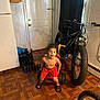 child, indoor, tricycle, bicycle, wooden_floor, door, curtain, red_shorts, black_slides, flooring, toy, playful, parquet_floor, stroller, bag, wall, home, person, young_child, casual