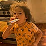 Jayden is registered to the contest to win money with this photo: child, curly_hair, yellow_shirt, dinosaur_print, snack, kitchen, stove, wooden_floor, pouch, indoor, person, food, casual_clothing, young, playful, home, curious, brown_hair, floor, cooking_area