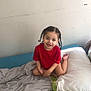 child, toddler, smiling, braided_hair, red_shirt, bed, blanket, pillow, indoor, wall, happy, person, young_child, casual, seated, home, cute, portrait, comfortable, relaxed