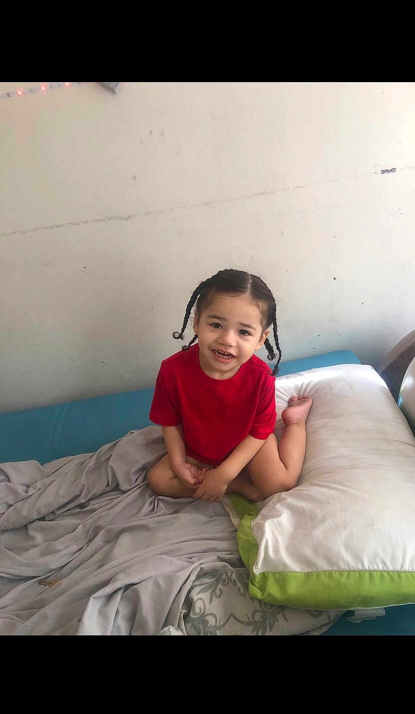 Jayden joined the competition — help win amazing prizes! child, toddler, smiling, braided_hair, red_shirt, bed, blanket, pillow, indoor, wall, happy, person, young_child, casual, seated, home, cute, portrait, comfortable, relaxed