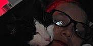 Luka is registered to the contest to win money with this photo: cat, person, glasses, face, black_and_white_cat, indoor, red_light, reflection, close_up, snuggling, pet, dark, cozy, affection, relaxing, feline, portrait, cuddle, animal, eyewear