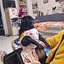 dog, pug, pet, dress, clothing, indoor, living_room, person, sitting, furniture, shelf, books, decorations, floor, carpet, slippers, yellow_jacket, black_dog, cute, animal