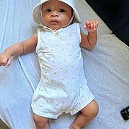 Maëylan a rejoint le concours — aidez-le/la à gagner de superbes lots ! baby, barefoot, child, cute, expression, face, hat, indoor, infant, light, looking_at_camera, lying_down, person, relaxed, remote_control, romper, skin, soft_surface, white_clothing, yellow_prints