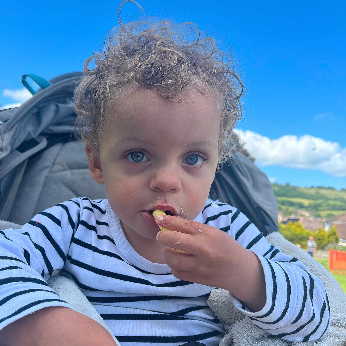 Amari is registered to the contest to win money with this photo: blanket, blue_eyes, casual_clothing, child, clouds, curly_hair, daylight, face, grass, hand, nature, outdoor, person, portrait, seated, sky, snack, striped_shirt, stroller, toddler