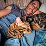 man, cat, tabby_cat, bed, blanket, blue_blanket, snuggling, relaxation, indoor, portrait, human_face, pet, sleeping_cat, comfort, cozy, resting, casual, home, feline, closeup