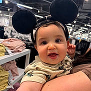 Enzo is registered to the contest to win money with this photo: baby, child, mouse_ears, headband, smiling, indoor, store, clothing, shelf, person, cute, face, holding, shopping, bright_lights, curious, toddler, apparel, human, portrait