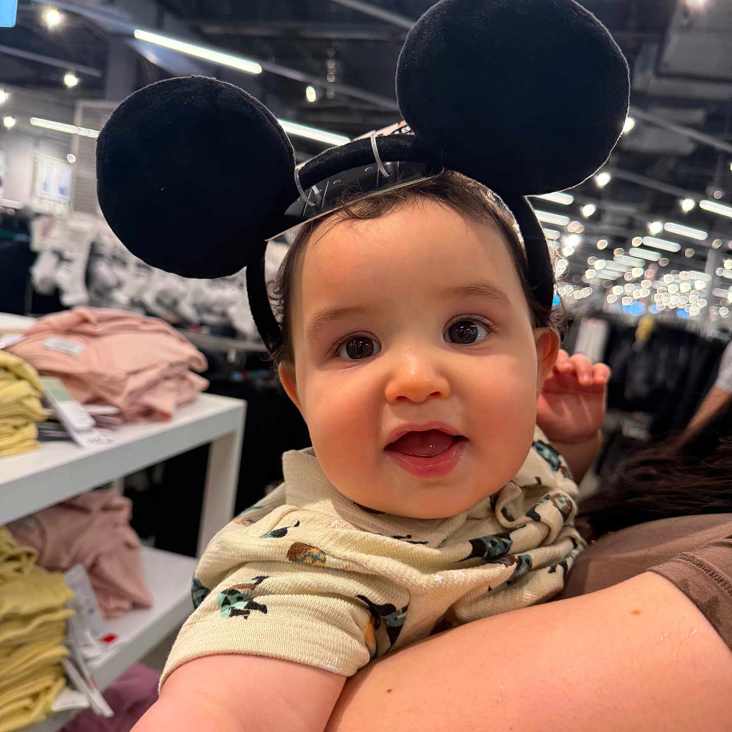 Enzo is registered to the contest to win money with this photo: baby, child, mouse_ears, headband, smiling, indoor, store, clothing, shelf, person, cute, face, holding, shopping, bright_lights, curious, toddler, apparel, human, portrait