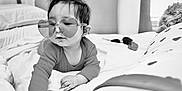 Matthew is registered to the contest to win money with this photo: baby, child, heart_shaped_glasses, glasses, bed, blanket, pillow, indoor, black_and_white, cute, infant, clothing, portrait, person, soft_texture, hair, young_child, lying_down, cozy, curious