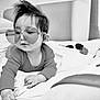 baby, child, heart_shaped_glasses, glasses, bed, blanket, pillow, indoor, black_and_white, cute, infant, clothing, portrait, person, soft_texture, hair, young_child, lying_down, cozy, curious
