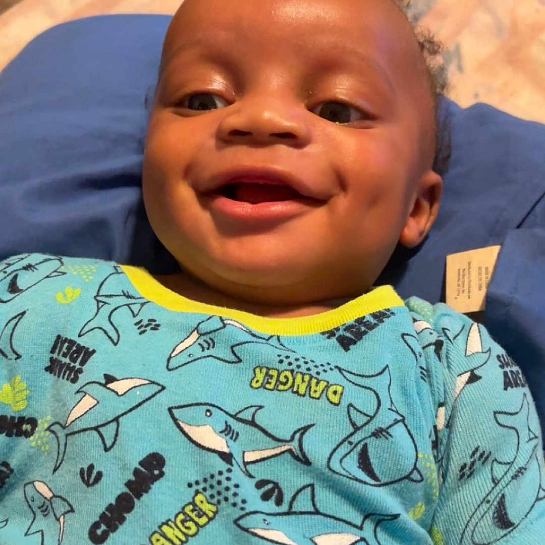 Mahari joined the competition — help win amazing prizes! baby, child, smiling, blue_clothing, shark_pattern, onesie, dimples, face, happy, infant, cute, person, portrait, indoor, cushion, fabric, textile, skin, head, lying_down