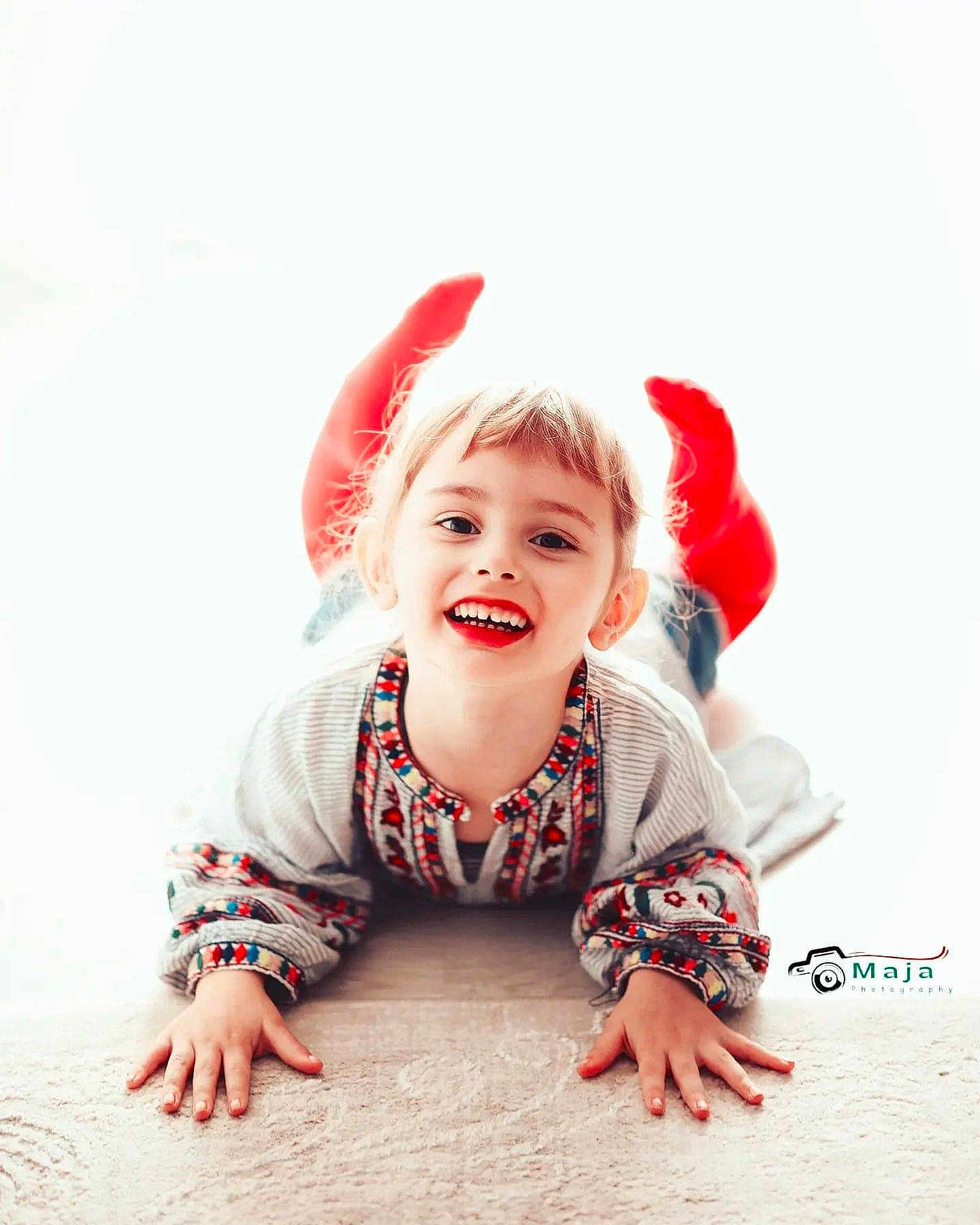 Maja a rejoint le concours — aidez-le/la à gagner de superbes lots ! child, smiling, red_socks, embroidered_shirt, lying_down, happy, face, hands, floor, portrait, cute, young, indoor, person, bright_background, casual_clothing, playful, joyful, childhood, light