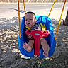 baby, infant, child, swing, playground, outdoor, smiling, happy, laughing, seated, safety_harness, blue_swing, red_safety_bar, rope, sunlight, sand, road, fence, trees, portrait