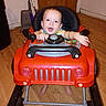baby, child, toy_car, walker, smiling, happy, indoor, portrait, face, steering_wheel, red, vehicle, playtime, sitting, bare_feet, wooden_floor, door, home, infant, looking_at_camera