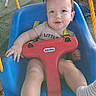 Lucas joined the competition — help win amazing prizes! baby, child, infant, swing, playground, outdoor, smiling, happy, portrait, face, eyes, brown_hair, blue_seat, red_safety_bar, yellow_rope, safety_harness, seated, play_equipment, adult_hand, daylight