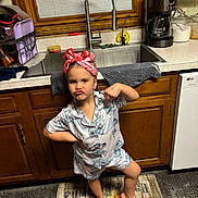 Phoenix is registered to the contest to win money with this photo: child, pajamas, pink_headscarf, barefoot, kitchen, sink, tap, wooden_cabinet, dish_towel, coffee_maker, rug, pumpkin, expression, pose, window, countertop, floor, appliance, smirk, playful