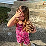 child, girl, pine_cone, pink_clothing, sneakers, outdoor, daylight, steps, house, concrete, gravel, smiling, playful, long_hair, hand, footwear, front_door, backpack, chalk, shadow