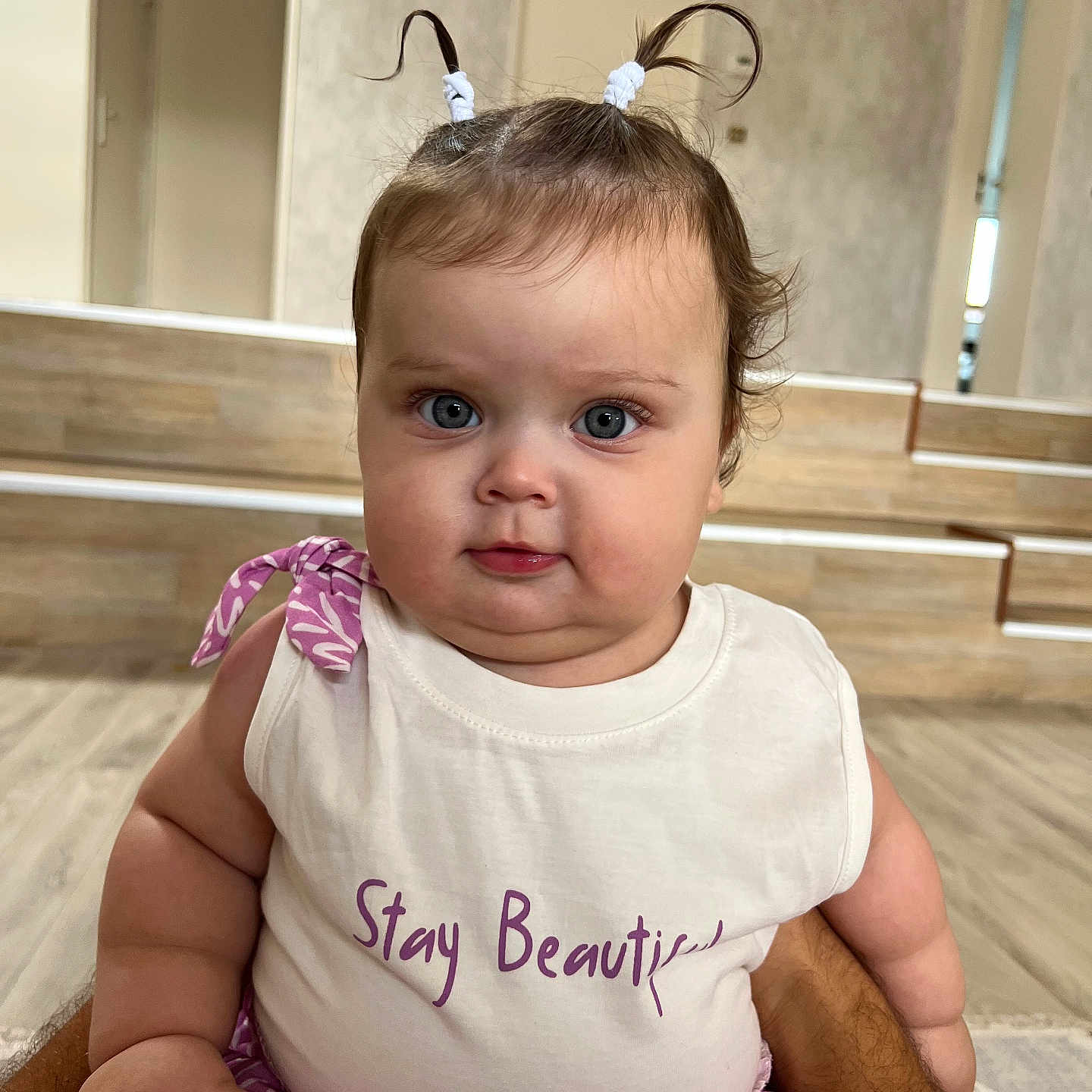Mira participe au concours pour gagner de l'argent avec cette photo : arms, baby, casual_clothing, child, chubby_cheeks, cute, expression, face, floor, hand, indoor, looking_at_camera, person, pigtails, portrait, purple_clothing, sitting, steps, white_top, wooden_floor