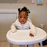 Janelle is registered to the contest to win money with this photo: toddler, child, high_chair, indoor, curly_hair, hair_ties, pajamas, white_clothing, face, eyes, hands, expression, floor, wall, door, electrical_outlet, thermostat, furniture, person, home