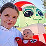 baby_doll, baby_yoda, child, christmas, face, festive, field, girl, holiday, holiday_decoration, hoodie, inflatable, outdoor, person, portrait, santa_hat, smile, sunny, toy, tree