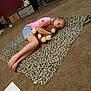 child, girl, stuffed_animal, blanket, leopard_print, carpet, indoor, casual_clothing, pink_shirt, denim_shorts, barefoot, relaxing, toy, floor, home, cozy, lying_down, paper, electronics, shelf