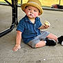 background, blue_shirt, casual, child, concrete_floor, cupcake, curious, face, fingers, footwear, frosting, hand, hat, outdoor, person, shade, shorts, sitting, table_leg, toddler