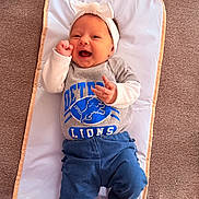 Emery is registered to the contest to win money with this photo: baby, infant, smiling, headband, bow, detroit_lions, shirt, blue_pants, white_socks, lying_down, padded_mat, carpet, happy, cheerful, cute, child, person, indoors, portrait, clothing