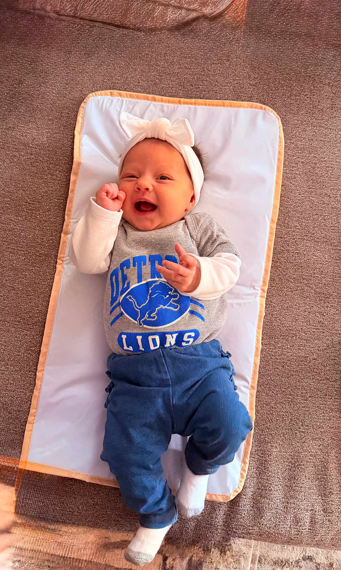 Emery is registered to the contest to win money with this photo: baby, infant, smiling, headband, bow, detroit_lions, shirt, blue_pants, white_socks, lying_down, padded_mat, carpet, happy, cheerful, cute, child, person, indoors, portrait, clothing
