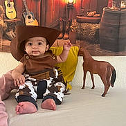 Zayan participe au concours pour gagner de l'argent avec cette photo : baby, child, cowboy_hat, toy_horse, adult, bed, yellow_pillow, western_theme, guitar, boots, plaid_pants, indoor, person, cute, smiling, costume, hat, foot, hand, face