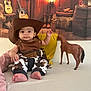 baby, child, cowboy_hat, toy_horse, adult, bed, yellow_pillow, western_theme, guitar, boots, plaid_pants, indoor, person, cute, smiling, costume, hat, foot, hand, face