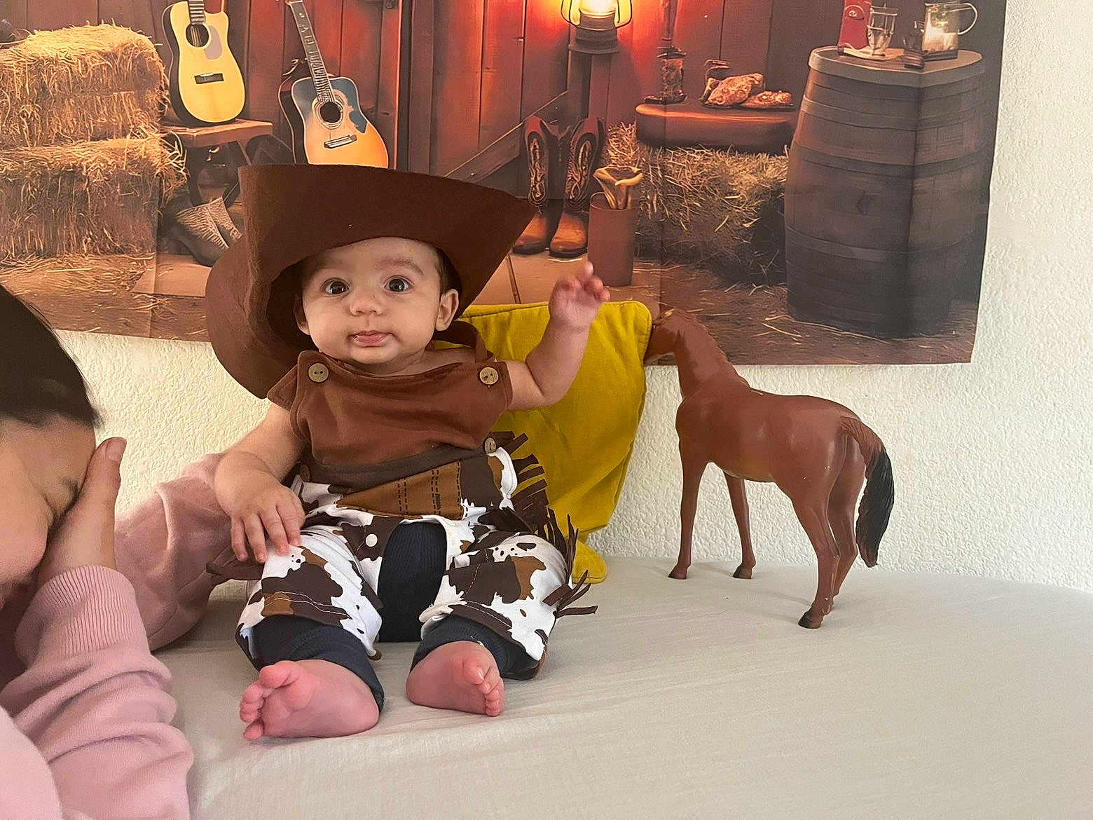 Zayan participe au concours pour gagner de l'argent avec cette photo : baby, child, cowboy_hat, toy_horse, adult, bed, yellow_pillow, western_theme, guitar, boots, plaid_pants, indoor, person, cute, smiling, costume, hat, foot, hand, face