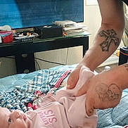 Sheryl is registered to the contest to win money with this photo: adult, baby, bed, blanket, box, changing_diaper, cords, gaze, glasses, hand, indoor, infant, onesie, patterned_blanket, pillow, portrait, skin, table, tattoo, television