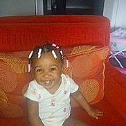 Caliyah joined the competition — help win amazing prizes! toddler, child, smiling, sitting, orange_couch, hair_beads, indoors, cushions, happy, person, baby, casual_clothing, home, cute, portrait, furniture, living_room, infant, seated, young_child