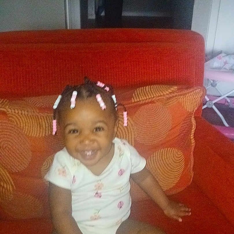 Caliyah joined the competition — help win amazing prizes! toddler, child, smiling, sitting, orange_couch, hair_beads, indoors, cushions, happy, person, baby, casual_clothing, home, cute, portrait, furniture, living_room, infant, seated, young_child