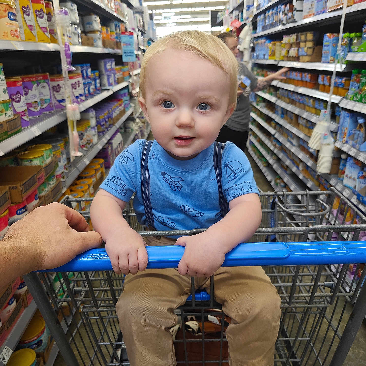 Zacharah is registered to the contest to win money with this photo: adult_hand, aisle, blonde_hair, blue_eyes, blue_shirt, casual_clothing, child, cute, grocery_store, holding_bar, indoor, person, products, retail, shelves, shopping, shopping_cart, supermarket, tan_pants, toddler
