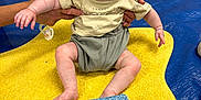 Jude is registered to the contest to win money with this photo: baby, child, green_mask, superhero_mask, yellow_mat, colorful_blocks, pacifier, adult_hands, indoor, floor, smiling, happy, cute, clothing, shorts, tshirt, toy, playtime, infant, portrait