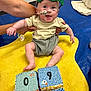 Jude is registered to the contest to win money with this photo: baby, child, green_mask, superhero_mask, yellow_mat, colorful_blocks, pacifier, adult_hands, indoor, floor, smiling, happy, cute, clothing, shorts, tshirt, toy, playtime, infant, portrait