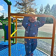 Anthony is registered to the contest to win money with this photo: toddler, child, playground, merry_go_round, snowsuit, yellow_bar, outdoor, happy, smiling, sunlight, trees, play_equipment, blue_floor, metal, park, daytime, person, clothing, baby, fun