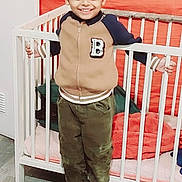 Maruthy participe au concours pour gagner de l'argent avec cette photo : bed, boy, casual_clothing, child, cozy, crib, furniture, grip, happy, indoors, jacket, orange_wall, pants, person, playful, shoes, smiling, standing, wooden_floor, young