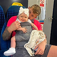 Novella is registered to the contest to win money with this photo: baby, man, baby_carrier, bow, tattoo, train_seat, train_interior, smile, white_shoes, knitted_blanket, pacifier, shorts, pink_shirt, window, door, sign, person, child, indoor, transport